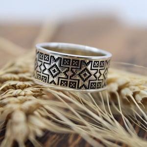 May include: A silver ring featuring a black and white geometric design. The ring is resting on a background of wheat stalks.