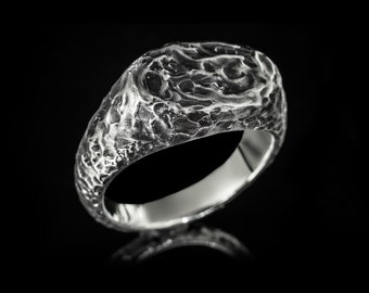 Puede incluir: Un anillo de plata con una superficie texturizada y rugosa. El anillo es ancho y tiene una parte superior redondeada.
