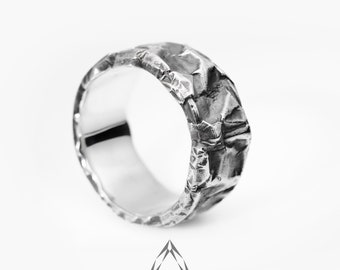 May include: A silver ring with a textured, crinkled surface. The ring has a wide band and a rough, uneven texture, creating a unique, organic appearance. The ring is set against a white background.