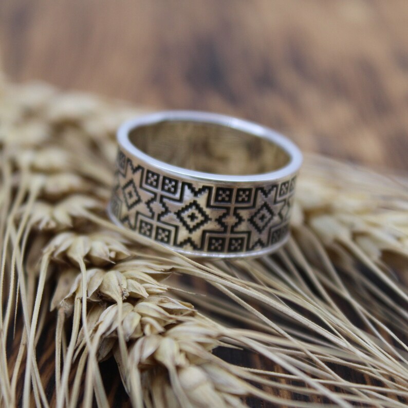 May include: A silver ring featuring a black geometric design. The ring is resting on a bed of wheat stalks.