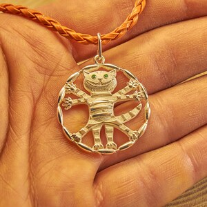 Peut inclure: Un collier avec un pendentif en argent rond et ajour&eacute; repr&eacute;sentant un chat stylis&eacute; aux yeux verts et un large sourire. Le chat est repr&eacute;sent&eacute; avec les bras et les jambes tendus, cr&eacute;ant une forme circulaire.