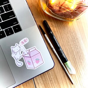 May include: A white cat sticker with a pink straw in its mouth, drinking from a pink and white carton of strawberry milk. The carton has Japanese text on it. The sticker is on a silver laptop.