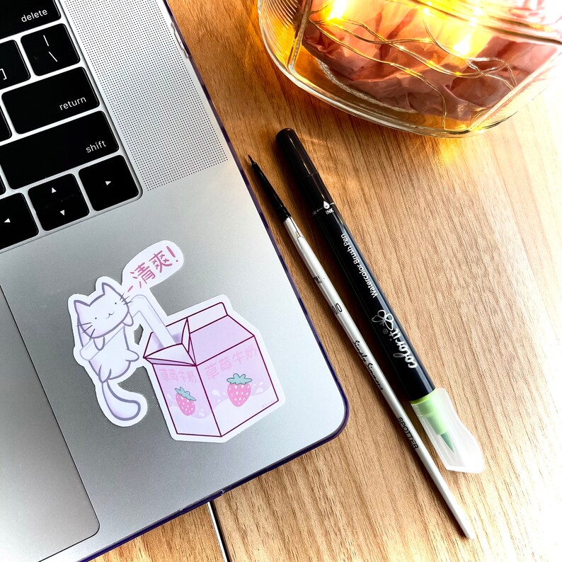 May include: A white cat sticker with a pink straw in its mouth, drinking from a pink and white carton of strawberry milk. The carton has Japanese text on it. The sticker is on a silver laptop.