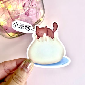 May include: A white and blue sticker with a cartoon brown cat peeking out of a white steamed bun. The sticker has the Chinese text "小笼猫" which translates to "Little Steamed Bun Cat".