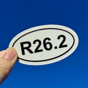 Może przedstawiać: Owalna naklejka z czarnym napisem "R26.2" na tle jasnoniebieskiego tła. Naklejka ma białe tło i czarną obwódkę. Tekst prawdopodobnie odnosi się do dystansu maratonu.