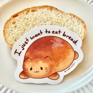 May include: A sticker depicting a cartoon cat resembling a loaf of bread, with the words "I just want to eat bread." The sticker is on a slice of bread, set against a white plate.