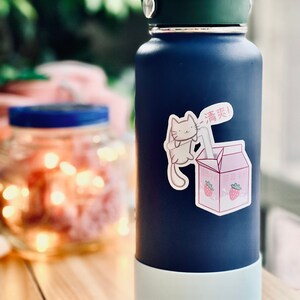 May include: A blue reusable water bottle with a white bottom and a sticker of a cartoon cat drinking from a carton of strawberry milk. The sticker says "Drink!" in Japanese.
