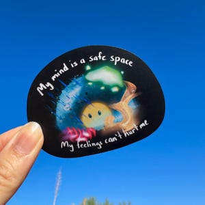 Könnte beinhalten: Ein schwarzer Aufkleber mit dem Text "My mind is a safe space" und "My feelings can't hurt me." Der Aufkleber zeigt eine Cartoon-Illustration eines gelben Charakters mit einem lächelnden Gesicht, umgeben von einem farbenfrohen, verträumten Design.