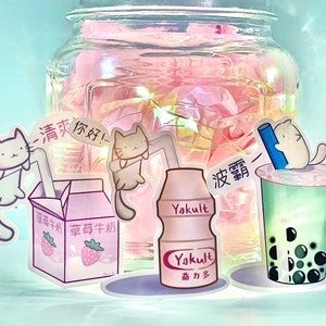 May include: A collection of cute cat stickers featuring a milk carton, a bottle of Yakult, and a boba tea cup. The stickers are on a pink background with a glass jar filled with pink tissue paper.