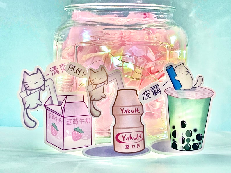 May include: A collection of cute cat stickers featuring a milk carton, a bottle of Yakult, and a boba tea cup. The stickers are on a pink background with a glass jar filled with pink tissue paper.