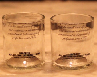 Könnte beinhalten: Zwei klare Glasbecher mit einer gedruckten Botschaft, die lautet: "in der Nähe der kleinen Stadt und setzt ein entschlossenes Engagement für das Streben nach Perfektion seit 1879 fort". Der Boden jedes Glases trägt die Aufschrift "IMPORTIERT 40% ALC/VOL. (80 PROOF) 750 ML".