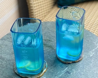 Könnte beinhalten: Zwei blaue Glasbecher gefüllt mit Eis und einer klaren Flüssigkeit, die auf runden grünen Untersetzern auf einem grünen Marmortisch stehen.