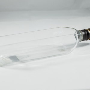 Puede incluir: Botella de vidrio transparente vac&iacute;a con una tapa de rosca negra y dorada.