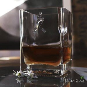 May include: A clear glass whiskey tumbler with a square shape and a walking man logo etched into the side. The glass is filled with amber-colored whiskey.