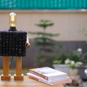 May include: A wooden robot lamp with a light bulb head made from computer keys. The robot has wooden arms and legs. The robot is sitting on a wooden table with a stack of books in front of it.