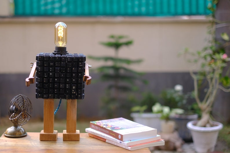 May include: A wooden robot lamp with a light bulb head made from computer keys. The robot has wooden arms and legs. The robot is sitting on a wooden table with a stack of books in front of it.