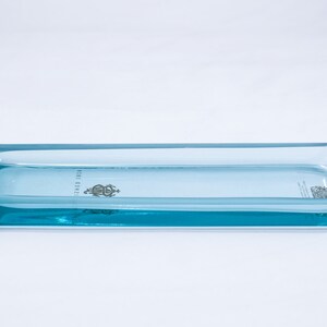 May include: A light blue glass tray with a raised edge and a slightly pointed end. The tray has a small, silver-colored logo in the centre.