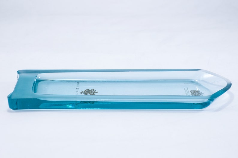 May include: A light blue glass tray with a raised edge and a slightly pointed end. The tray has a small, silver-colored logo in the centre.