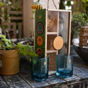 May include: Two square, teal-colored drinking glasses with gold lettering and a gold emblem. A wooden box with a bottle, decorative straw, and a "Hand Made" medallion is behind the glasses. A decorative bee and plants are in the background.