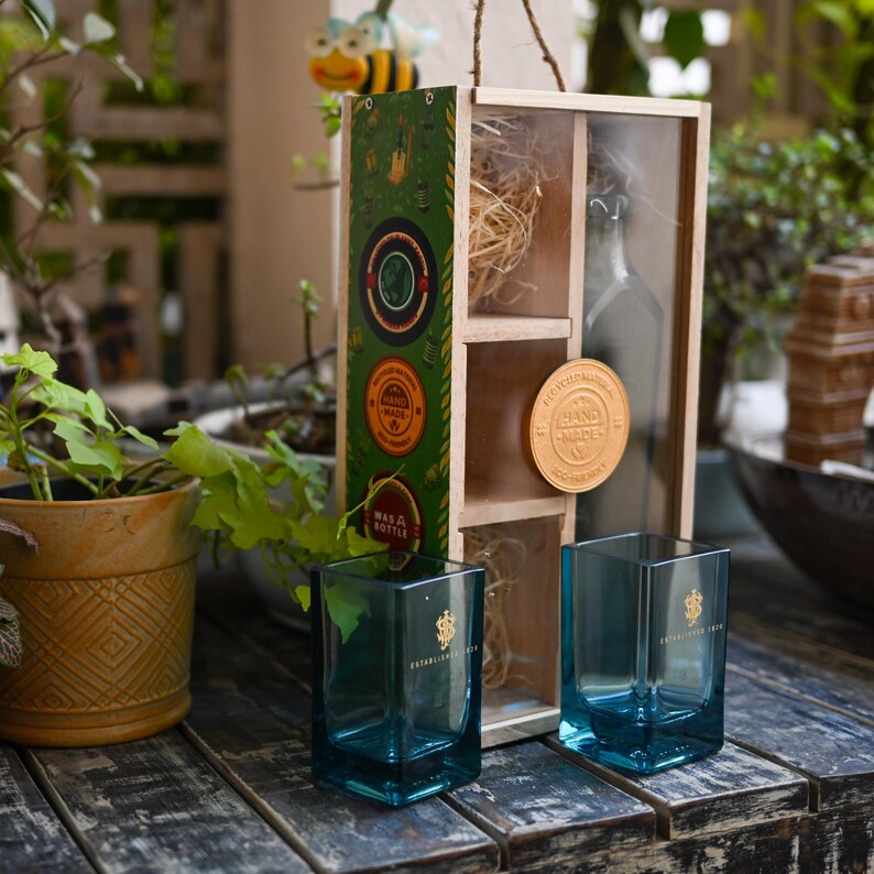May include: Two square, teal-colored drinking glasses with gold lettering and a gold emblem. A wooden box with a bottle, decorative straw, and a "Hand Made" medallion is behind the glasses. A decorative bee and plants are in the background.