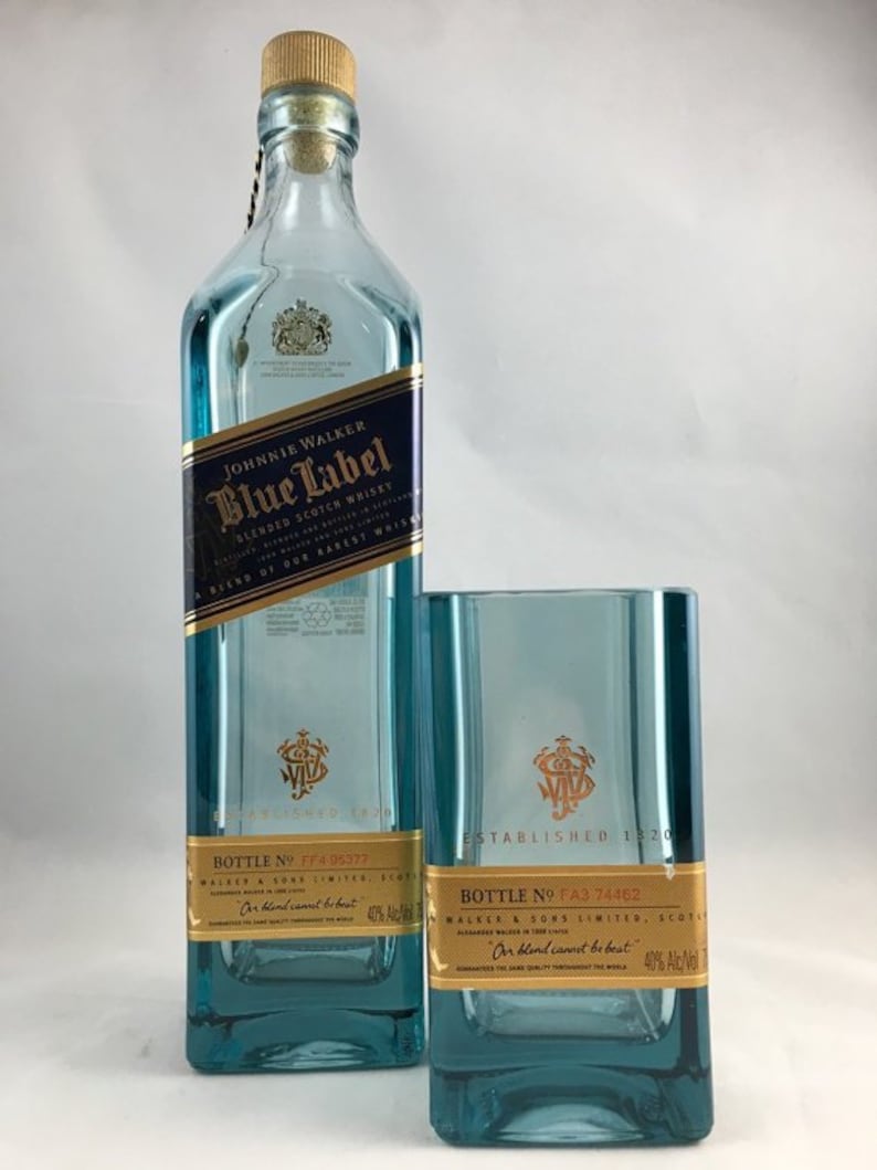 May include: A blue-tinted glass bottle of Johnnie Walker Blue Label blended scotch whisky with a matching glass. The bottle has a gold label and a wooden cork. The glass has a gold emblem and the text "ESTABLISHED 1820".