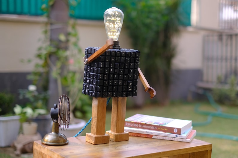May include: A lamp made from a computer keyboard, with wooden legs and arms, and a light bulb on top. The lamp is sitting on a wooden table with a few books stacked on top of it.