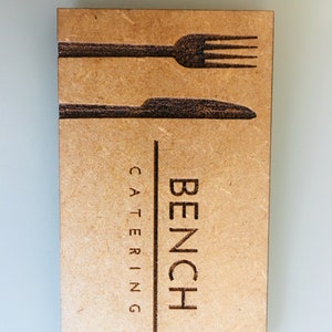 Op de afbeelding: Een bruine houten visitekaartje met de tekst "BENCH CATERING" en een silhouet van een vork en mes.