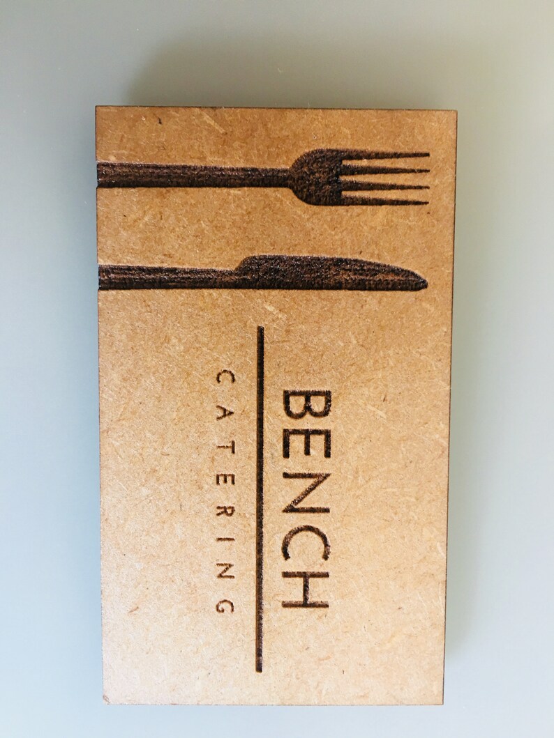 Op de afbeelding: Een bruine houten visitekaartje met de tekst "BENCH CATERING" en een silhouet van een vork en mes.