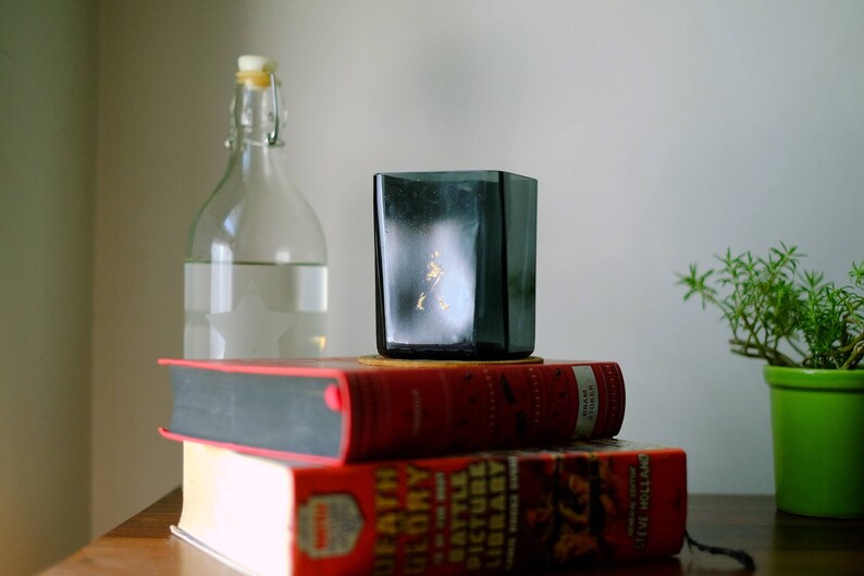 K&ouml;nnte beinhalten: Ein Stapel aus zwei roten Hardcover-B&uuml;chern mit einer durchsichtigen Glasflasche und einer dunkelgrauen Glas-Tasse darauf. Rechts im Bild steht eine kleine gr&uuml;ne Topfpflanze.