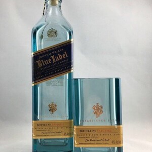 Puede incluir: Una botella de vidrio azul de whisky escoc&eacute;s Johnnie Walker Blue Label con una etiqueta dorada. La botella est&aacute; junto a un vaso de vidrio azul con una etiqueta dorada que dice "Establecido en 1820".