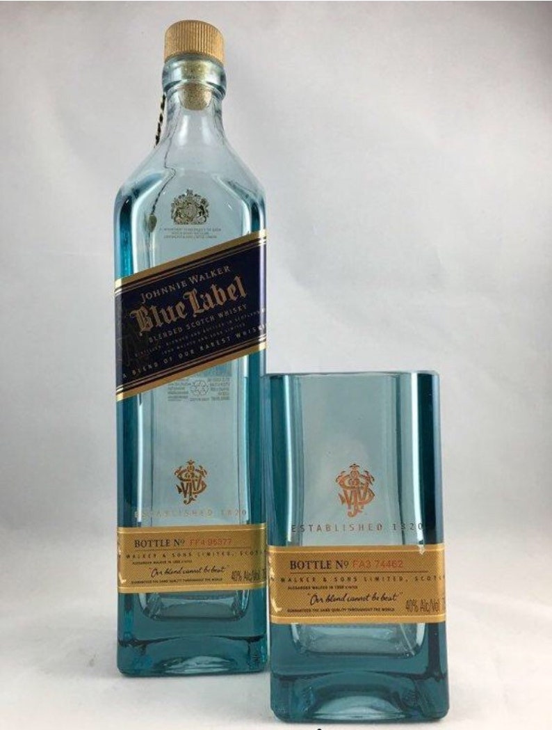 Puede incluir: Una botella de vidrio azul de whisky escoc&eacute;s Johnnie Walker Blue Label con una etiqueta dorada. La botella est&aacute; junto a un vaso de vidrio azul con una etiqueta dorada que dice "Establecido en 1820".