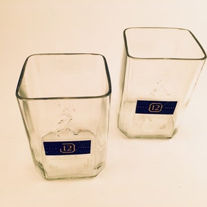 Op de afbeelding: Twee vierkante flessen van helder glas met een blauw etiket waarop "12 Years" staat en een nummer "12" in een cirkel.