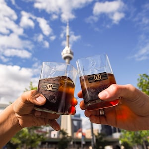 Op de afbeelding: Twee heldere glazen tumblers gevuld met een donker amberkleurige vloeistof, opgeheven in een toast. De glazen hebben een gouden label met het nummer "12" erop. De achtergrond is een wazig stadsbeeld met een hoge toren in de verte.