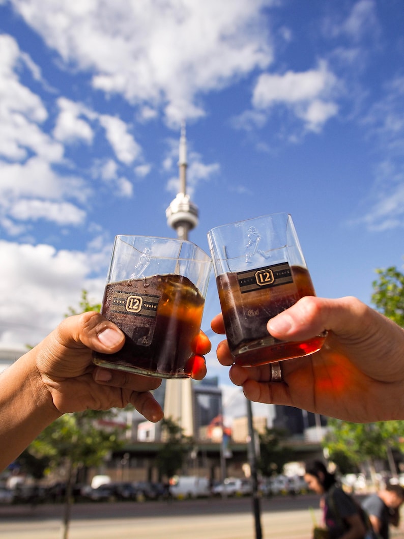 Op de afbeelding: Twee heldere glazen tumblers gevuld met een donker amberkleurige vloeistof, opgeheven in een toast. De glazen hebben een gouden label met het nummer "12" erop. De achtergrond is een wazig stadsbeeld met een hoge toren in de verte.