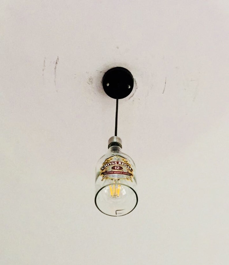 Peut inclure: Une bouteille en verre transparente avec une &eacute;tiquette Chivas Regal 12 est suspendue au plafond comme un luminaire. La bouteille est vide et contient une ampoule.