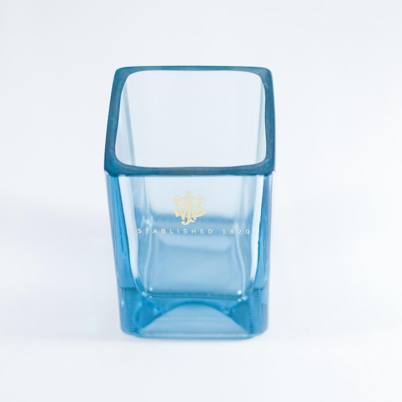 May include: A clear, light blue glass tumbler with a square top and a slightly tapered base. The rim and edges are a darker shade of blue. The glass features a gold emblem and the text "ESTABLISHED 1870".