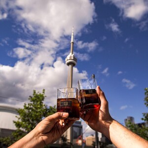 Op de afbeelding: Twee heldere glazen tumblers gevuld met een donkerbruine vloeistof, elk met een label met de tekst "12" en een gouden ontwerp. De glazen worden in een toast gehouden tegen een achtergrond van een blauwe lucht met witte wolken en een hoge toren in de verte.