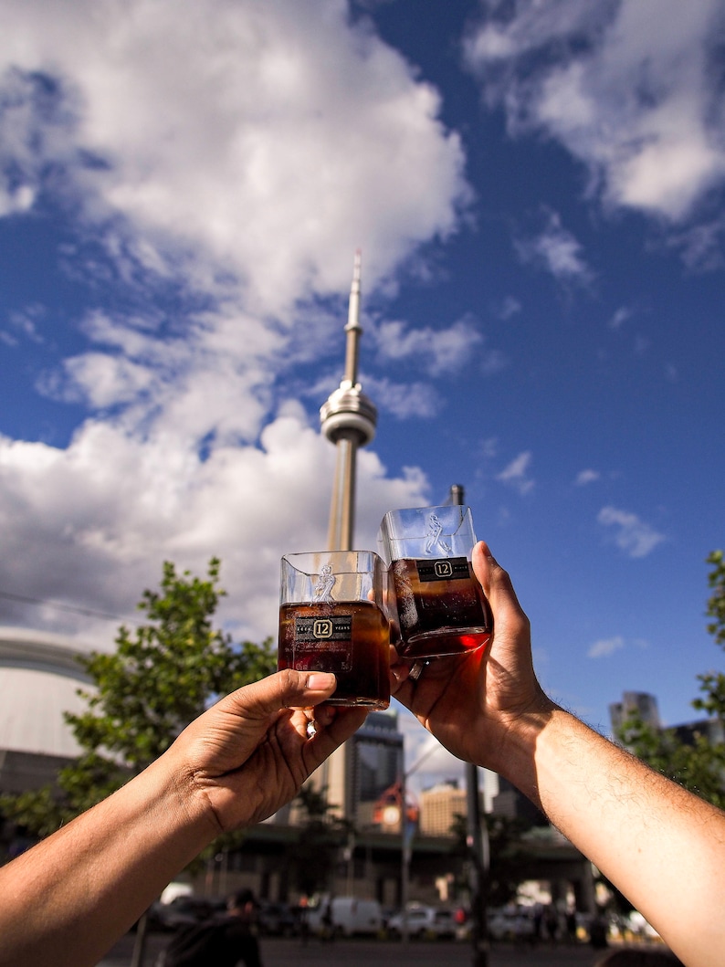 Op de afbeelding: Twee heldere glazen tumblers gevuld met een donkerbruine vloeistof, elk met een label met de tekst "12" en een gouden ontwerp. De glazen worden in een toast gehouden tegen een achtergrond van een blauwe lucht met witte wolken en een hoge toren in de verte.