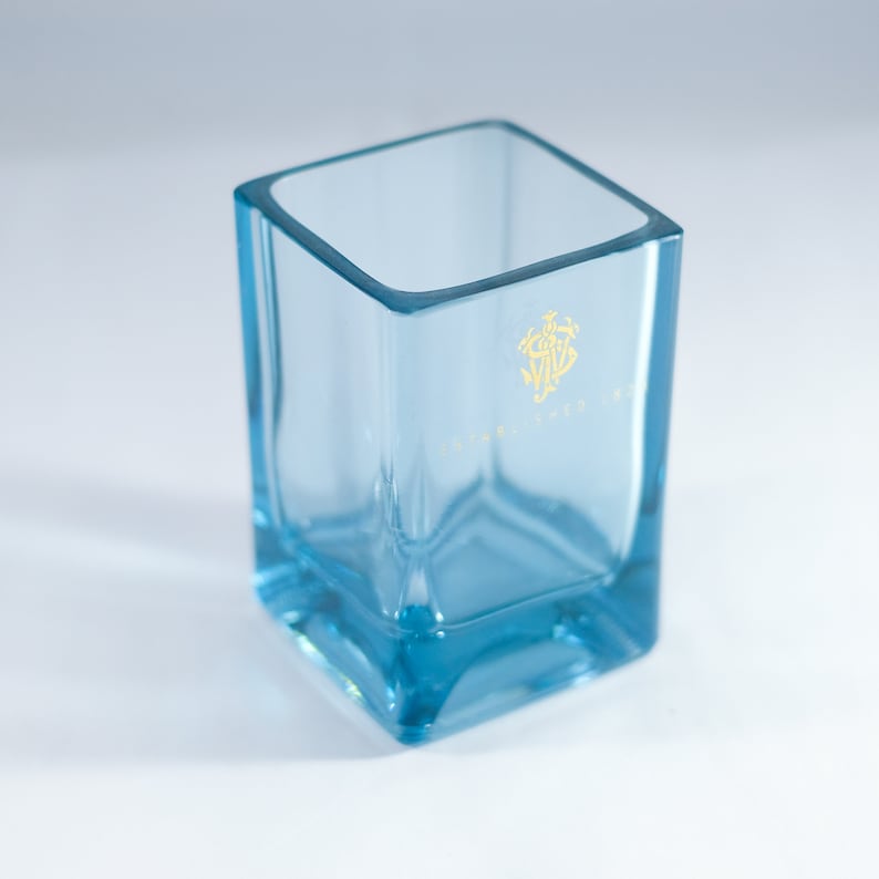 Puede incluir: Un vaso cuadrado de vidrio transparente con un tono azul claro. El vaso tiene un logotipo dorado en la parte delantera que dice "The Glenlivet".
