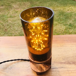 Könnte beinhalten: Eine handgefertigte Tischlampe mit einem bernsteinfarbenen Glasschirm und einem Holzsockel. Die Lampe hat eine dekorative Glühbirne mit kleinen, hellen Lichtern. Die Lampe ist mit einem schwarzen, geflochtenen Kabel angeschlossen. Die Lampe steht auf einem Holztisch.