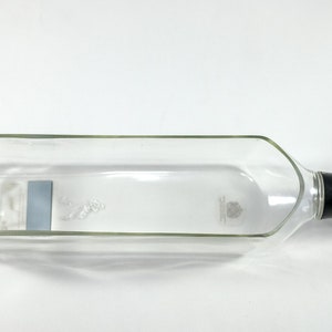 Puede incluir: Una botella de vidrio transparente vac&iacute;a con una tapa negra y dorada. La botella tiene una etiqueta en el lateral y est&aacute; tumbada de lado.