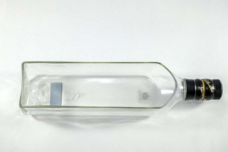 Puede incluir: Una botella de vidrio transparente vac&iacute;a con una tapa negra y dorada. La botella tiene una etiqueta en el lateral y est&aacute; tumbada de lado.