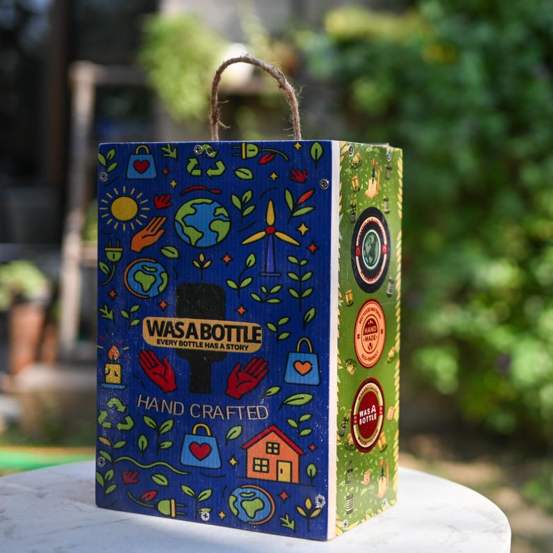 May include: A wooden box with a rope handle, featuring a blue front panel with colorful illustrations of the earth, a sun, and a windmill. The text "WAS A BOTTLE" and "HAND CRAFTED" are visible. The side panel is green with circular designs.