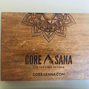 Op de afbeelding: Een houten bord met een mandala-ontwerp en de tekst "CORE ASANA" in zwarte letters. Onder de tekst staat de zin "FOR THE LOVE OF YOGA" en het websiteadres "COREASANA.COM".