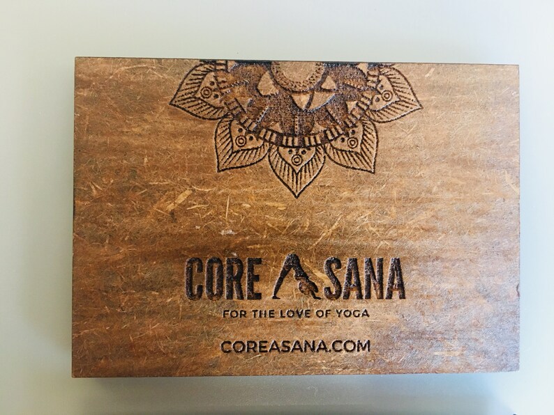 Op de afbeelding: Een houten bord met een mandala-ontwerp en de tekst "CORE ASANA" in zwarte letters. Onder de tekst staat de zin "FOR THE LOVE OF YOGA" en het websiteadres "COREASANA.COM".
