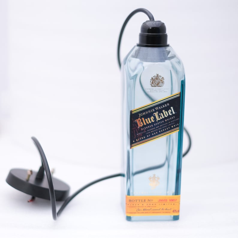 May include: A clear glass bottle with a black cord and plug. The bottle is labelled "Johnnie Walker Blue Label Blended Scotch Whisky A Blend of Our Rarest Whiskies". The bottle has a gold label with the text "Bottle No. 1443 1987", "Blended & Bottled", and "For blended Scotch whisky".