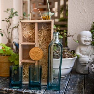 May include: A clear, rectangular glass bottle and two square glasses with a teal tint. A wooden box with a handle and a gold medallion with the words "HAND MADE" are also present. The items are arranged on a wooden surface.