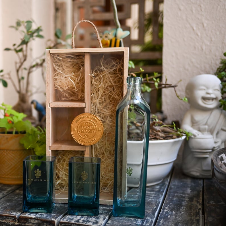 May include: A clear, rectangular glass bottle and two square glasses with a teal tint. A wooden box with a handle and a gold medallion with the words "HAND MADE" are also present. The items are arranged on a wooden surface.