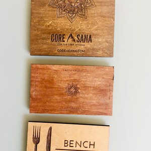 Op de afbeelding: Drie houten visitekaartjes met lasergegraveerde ontwerpen. De bovenste kaart is bruin met een mandala-ontwerp en de tekst "CORE ASANA FOR THE LOVE OF YOGA COREASANA.COM". De middelste kaart is roodbruin met een ster-ontwerp en de tekst "VASHU CHUGH 647-720-6219 VASHUYOGABRITY.COM". De onderste kaart is lichtbruin met een vork- en mesontwerp en de tekst "BENCH CATERING".