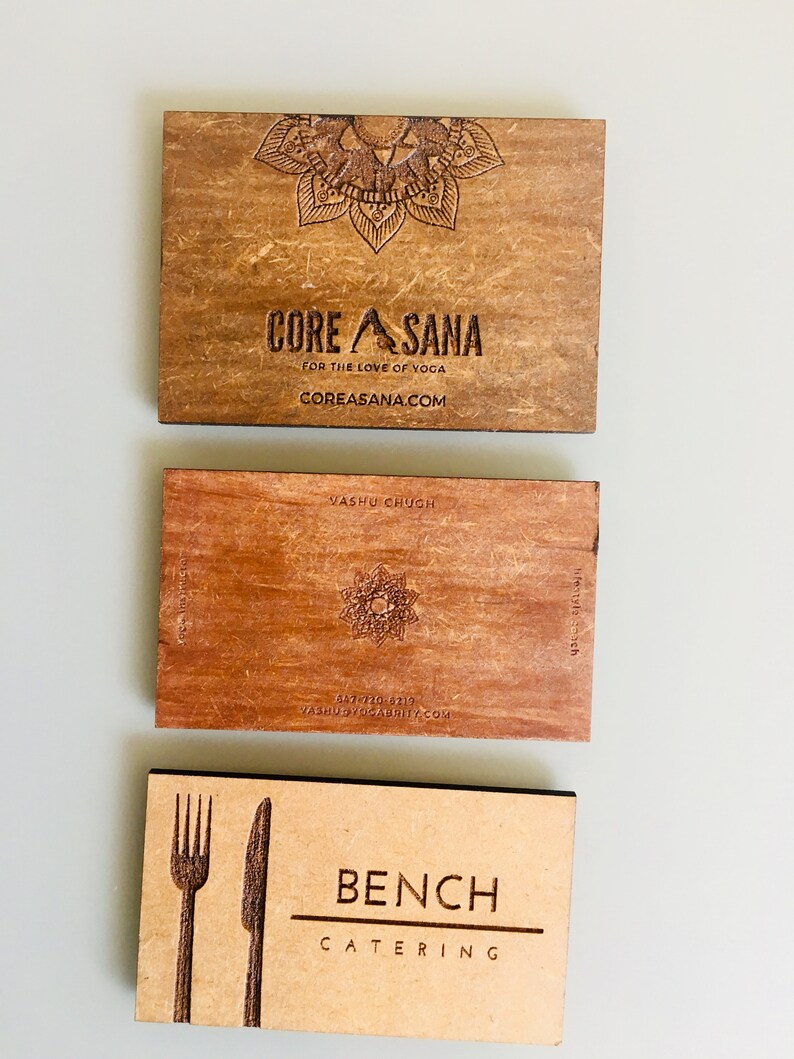 Op de afbeelding: Drie houten visitekaartjes met lasergegraveerde ontwerpen. De bovenste kaart is bruin met een mandala-ontwerp en de tekst "CORE ASANA FOR THE LOVE OF YOGA COREASANA.COM". De middelste kaart is roodbruin met een ster-ontwerp en de tekst "VASHU CHUGH 647-720-6219 VASHUYOGABRITY.COM". De onderste kaart is lichtbruin met een vork- en mesontwerp en de tekst "BENCH CATERING".